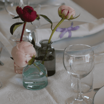 Ein festlich gedeckter Tisch mit weißem Porzellan und mehreren Gläsern. Dekoriert ist der Tisch mit dunkelroten und roséfarbenen Pfingstrosen und Stoffservietten mit lila Schleife (Foto: Bildehagen/Getty Images via Canva)